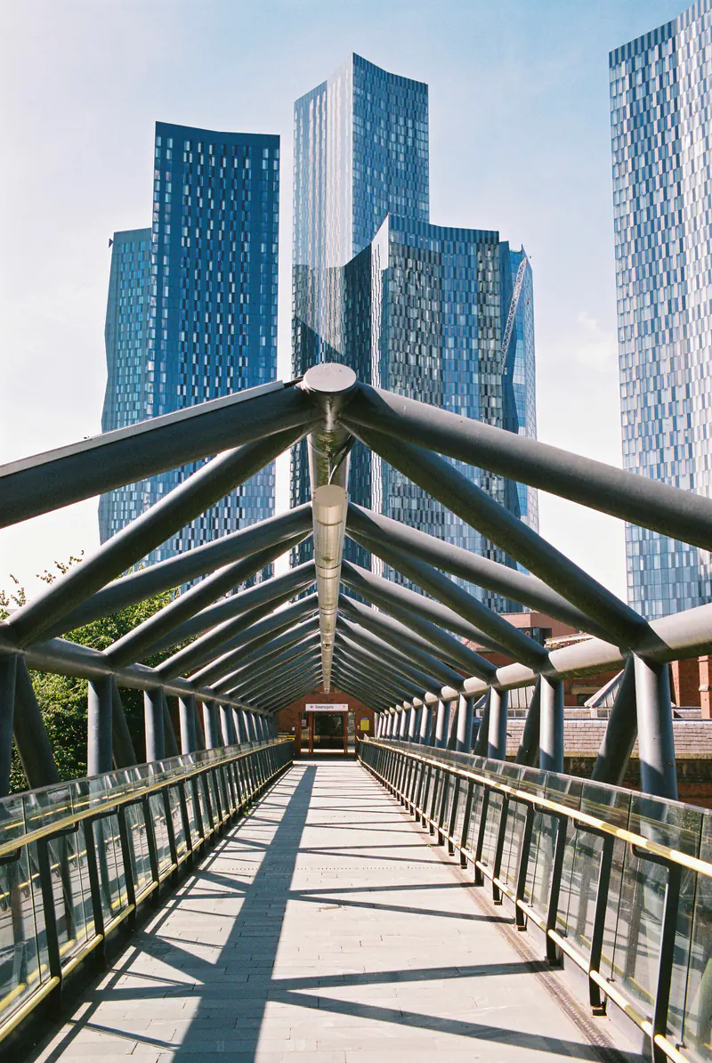 A covered steel footbridge leading toward the blue glass towers of Deansgate Square in Manchester.