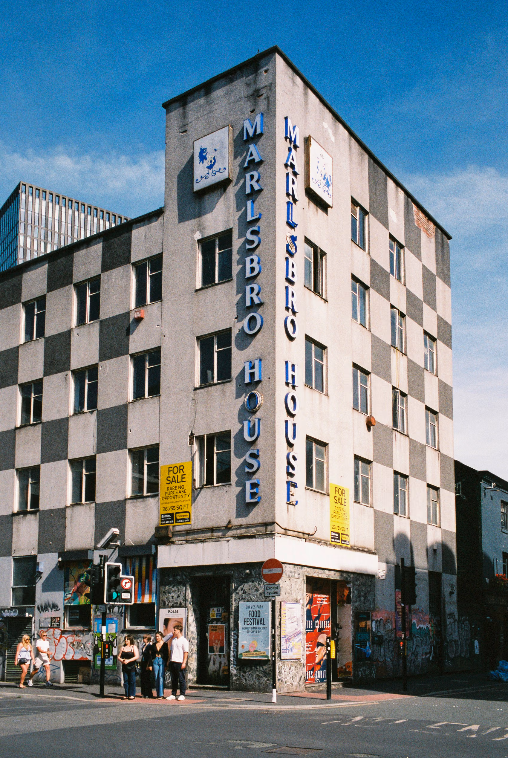 The grey-and-white checkerboard facade of Marlsbro House, a corner building in Manchester, against a clear blue sky.