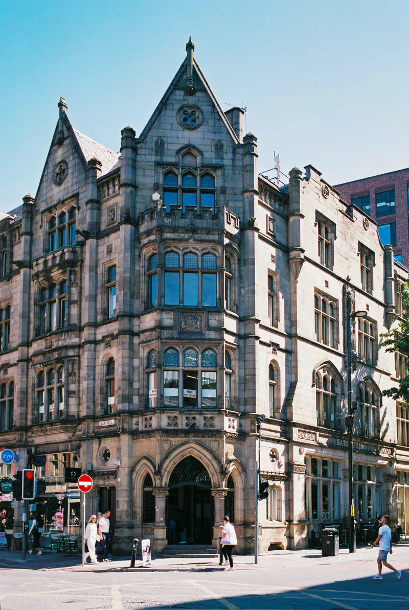 An ornate Victorian Gothic stone building with pointed gables on a Manchester street corner under a blue sky.