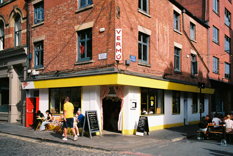 A red-brick corner building in Manchester housing Vermu, a bar with a yellow-painted front and people at outdoor tables.