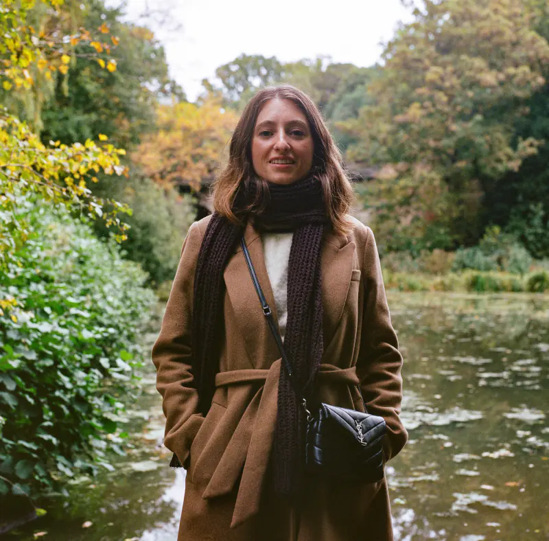 A woman wearing a brown coat and scarf is standing in front of a body of water.