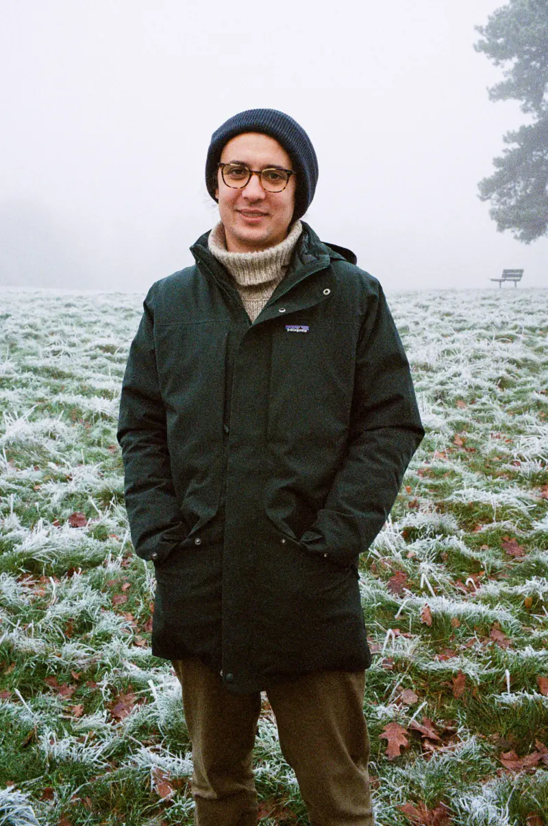 A man wearing a black jacket and glasses stands in a field.