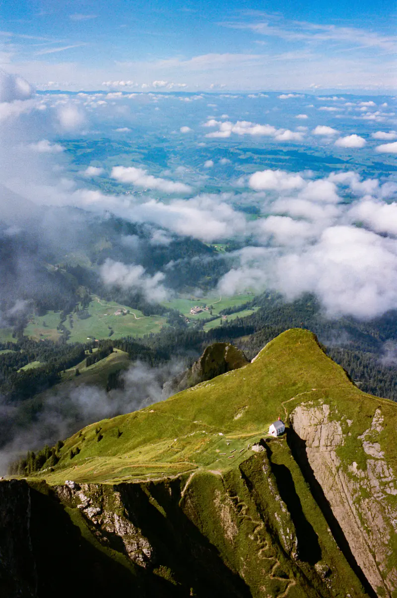 A small white chapel on a steep grassy alpine ridge, with clouds drifting over the green valley far below.