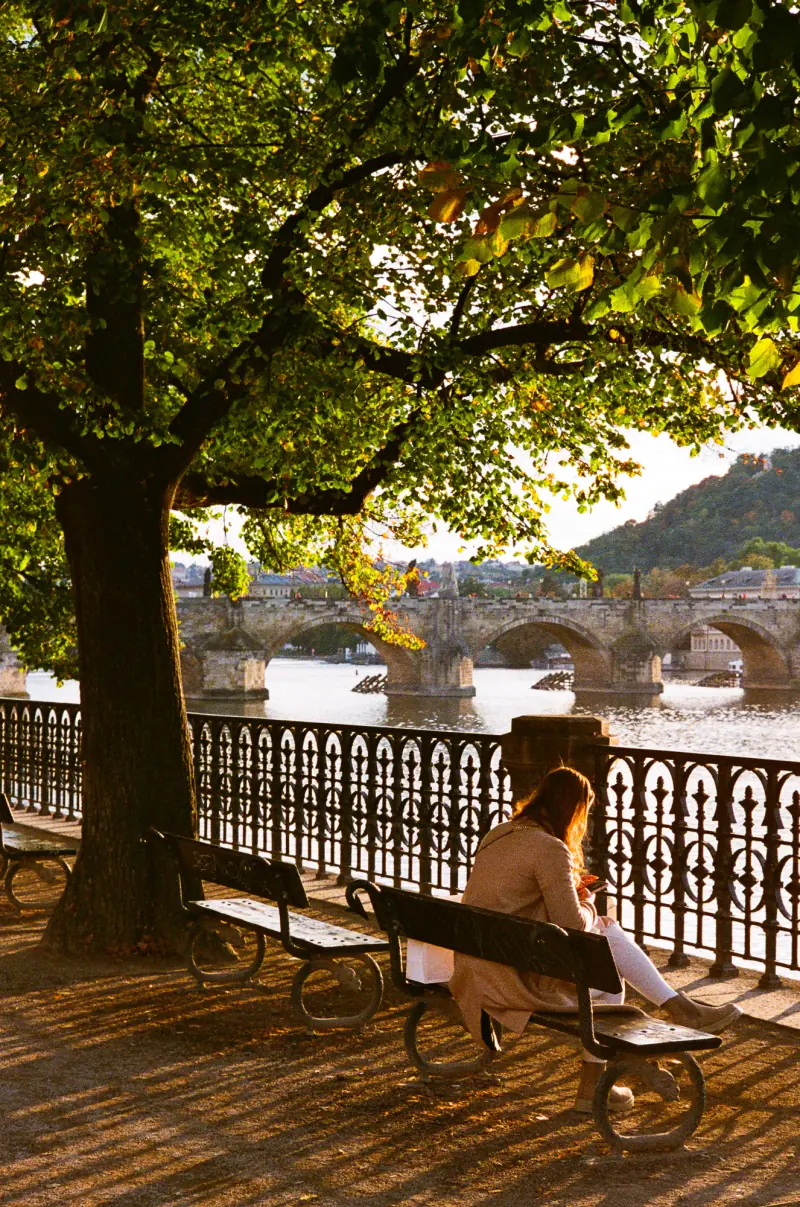 A woman sitting on a bench by a river with a bridge in the background.