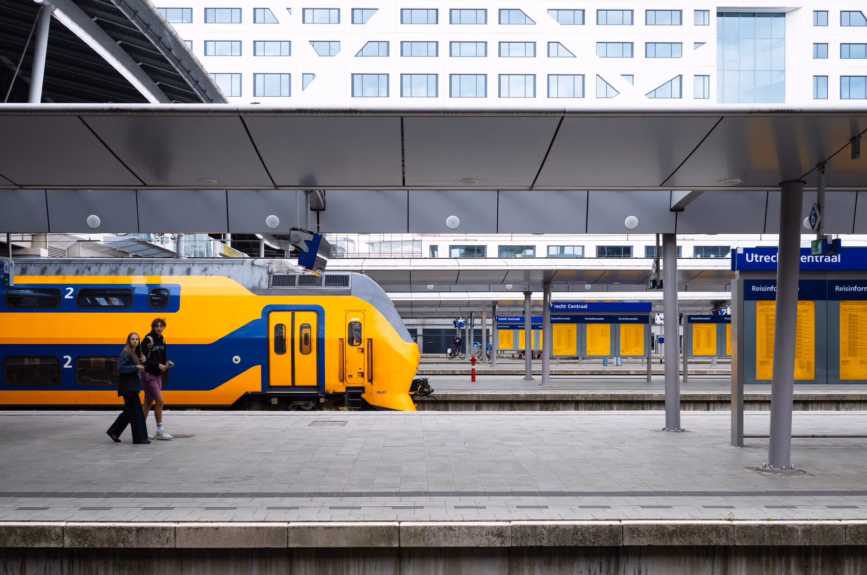 A yellow-and-blue Dutch double-decker train at a platform at Utrecht Centraal station, two people walking alongside it.